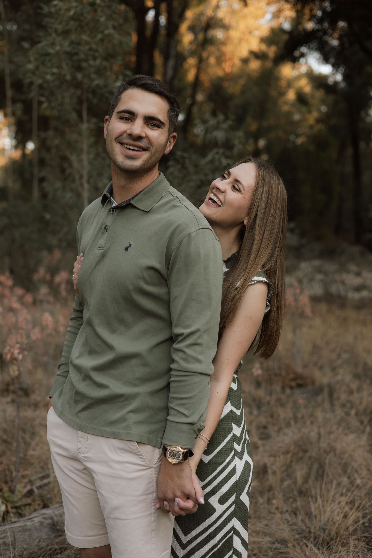 A smiling man in an olive polo holds hands with a laughing woman leaning on his shoulder in a golden autumn field.