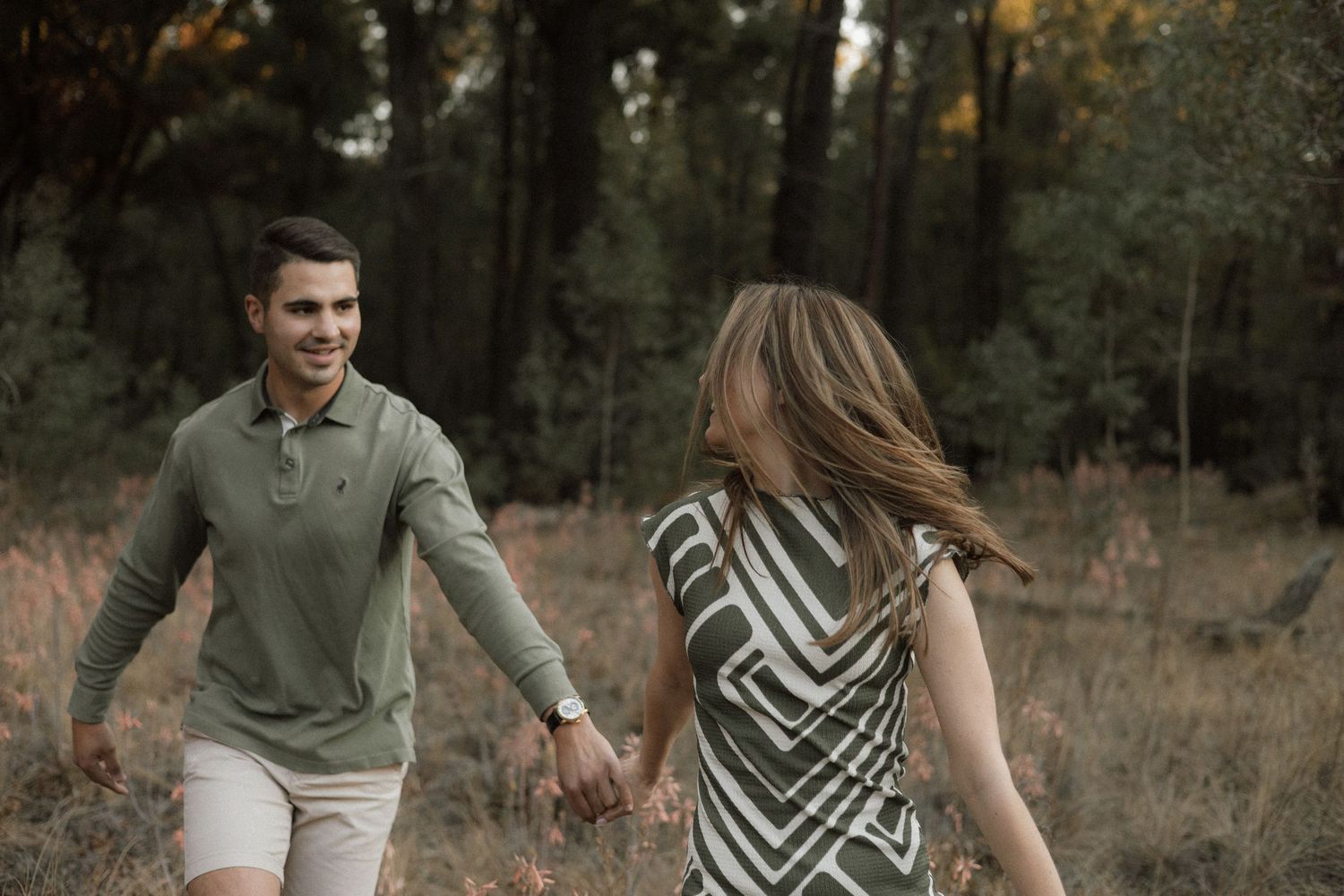 Smiling couple holds hands and walks through a golden grassy forest clearing at sunset during an outdoor engagement session.