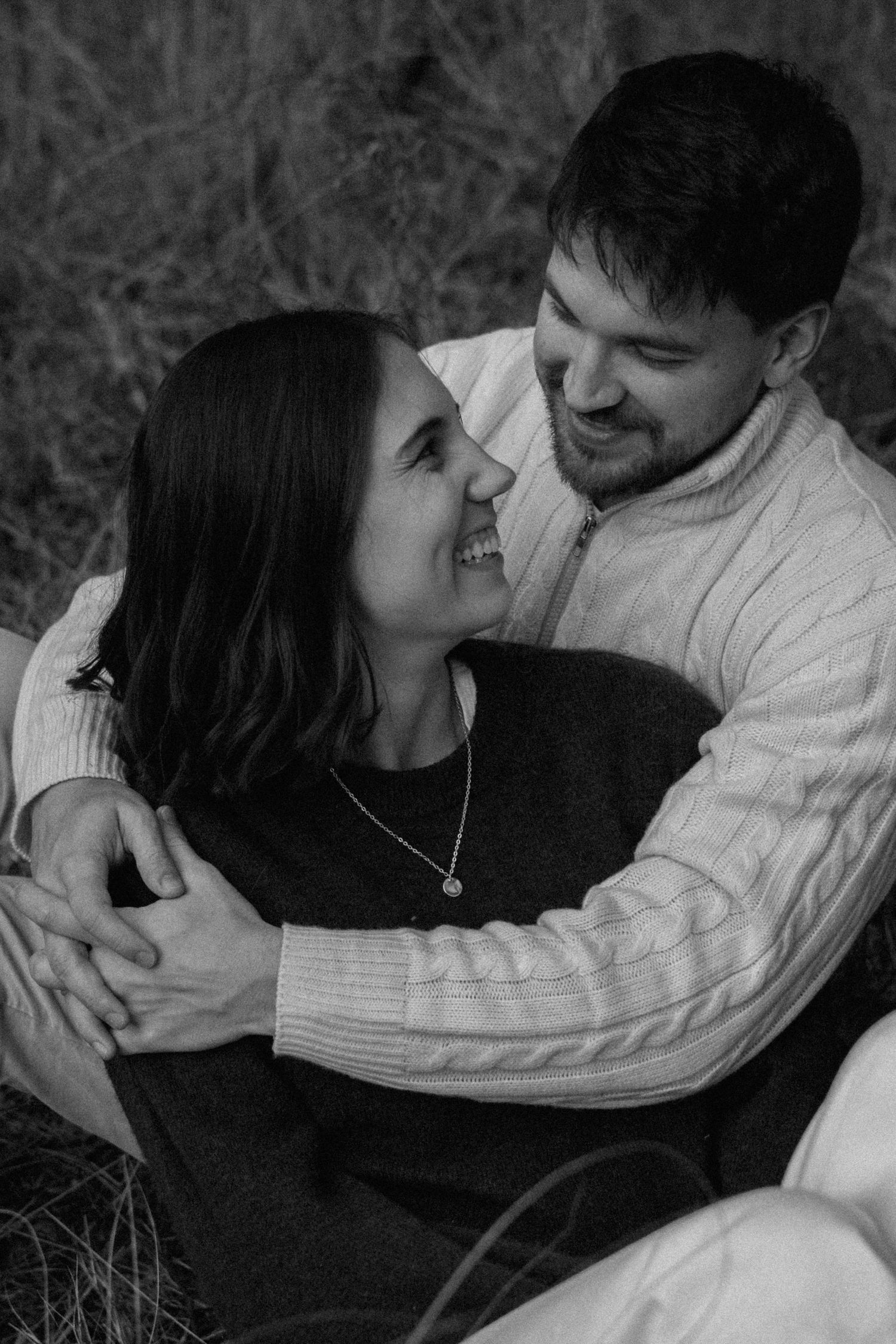 Black and white portrait of a couple sitting in tall grass, embracing and smiling warmly at each other.