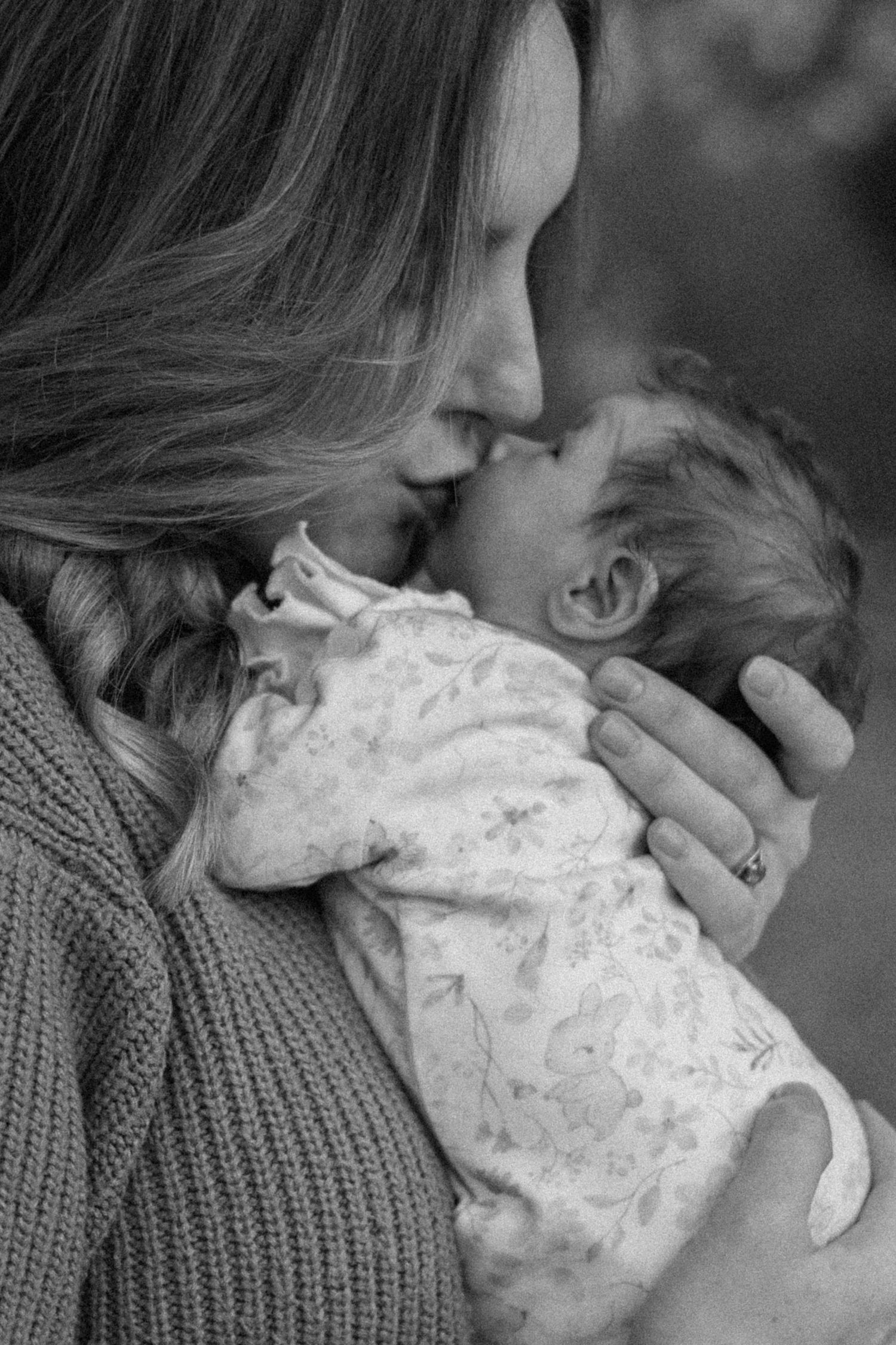 Black and white photo of a mother kissing her newborn baby, who is wrapped in a floral outfit, in a tender family portrait.