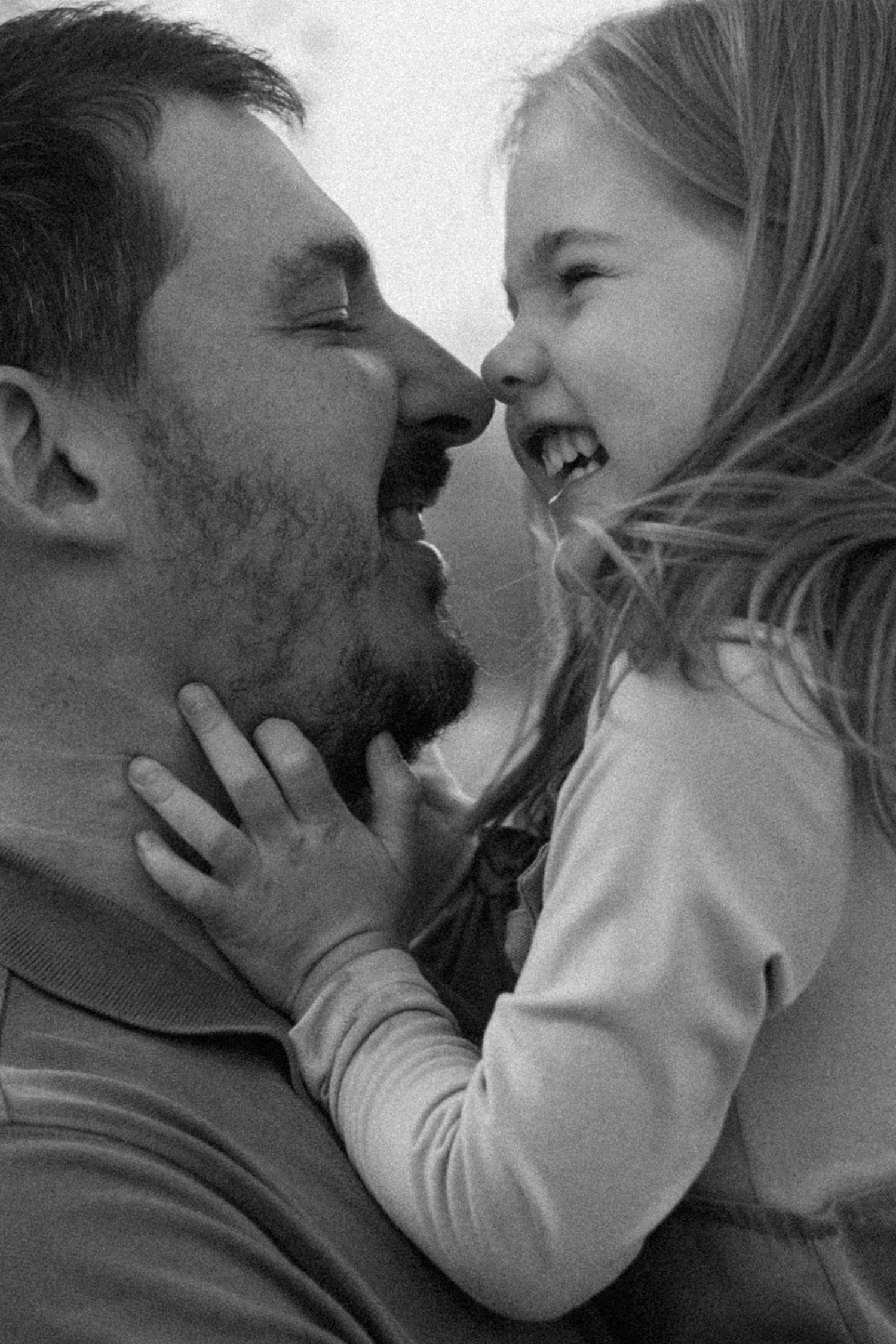 Black and white portrait of a smiling father and young daughter touching noses, her small hand resting gently on his bearded chin.