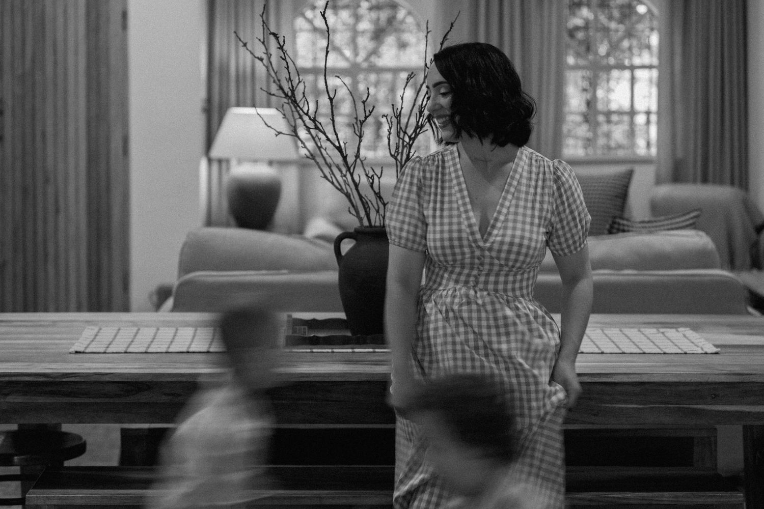 Black and white photo of a woman in a checkered dress leaning on a wooden table in a cozy living room as blurred children run past.